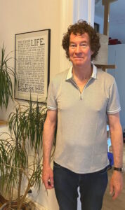 Photo of Richard Killian who has worked at PSU, Centre County Government. Background of a sign that tells how to live a good life with Richard Killian in the front standing in front of a plant.